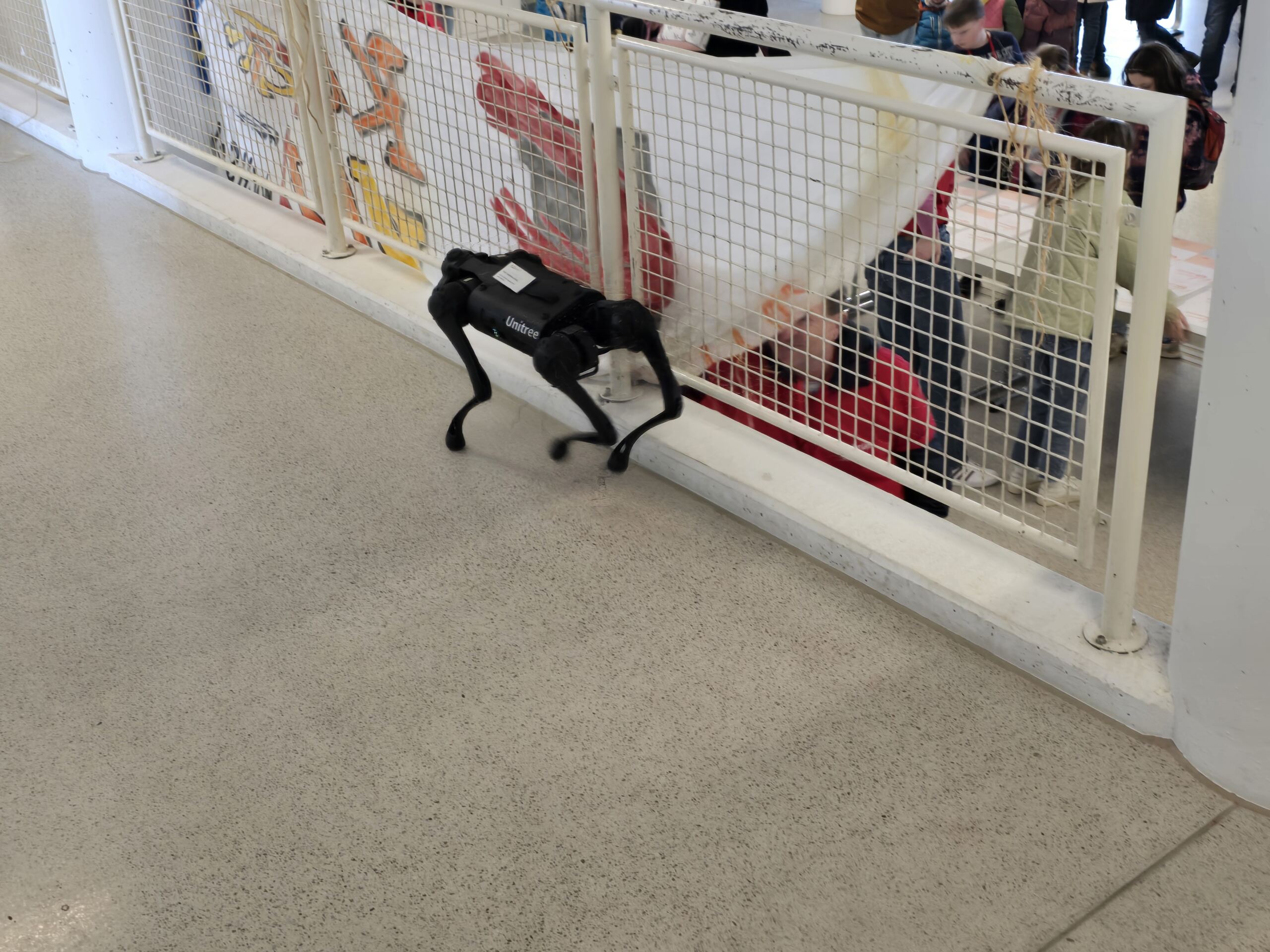 Ein Roboterhund spaziert durch einen eingezäunten Bereich, in dem sich Kinder und Erwachsene treffen, um mit farbenfrohen Kunstwerken zu interagieren. Die Inneneinrichtung, vermutlich Teil einer Kinder-Uni-Veranstaltung, ist mit glatten Böden und weißen Geländer ausgestattet. at Hochschule Coburg