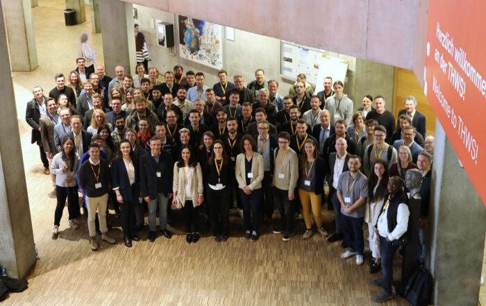 Eine große Gruppe von Menschen posiert für ein Foto in einer geräumigen Halle des NISys Promotionszentrums. Die meisten tragen Lanyards und lächeln. Das Ambiente ist modern und industriell gestaltet, mit Betonelementen und einem Forschungsbanner an der Wand. at Hochschule Coburg