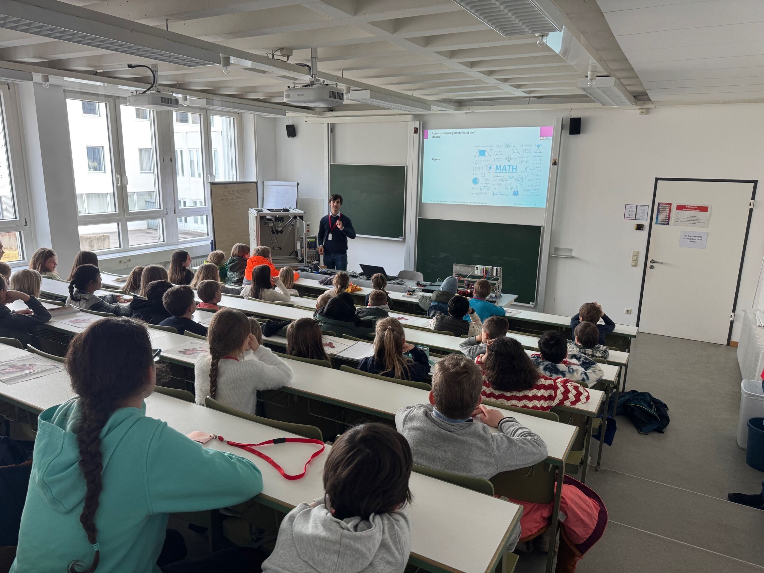 Ein Dozent steht vor einem Universitätshörsaal und spricht zu den Studierenden, die während einer speziellen Kinder-Uni-Veranstaltung in Reihen sitzen. Auf einer großen Leinwand hinter dem Dozenten läuft eine Präsentationsfolie mit Bildern vom jüngsten Jubiläum der Universität. Der Raum ist hell und hat Fenster an einer Seite. at Hochschule Coburg