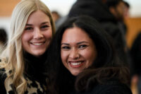 Two women smiling and posing together indoors. One has long blonde hair, and the other has long dark hair and a nose ring. Both are wearing black tops. The background is softly blurred with other people present. at Hochschule Coburg