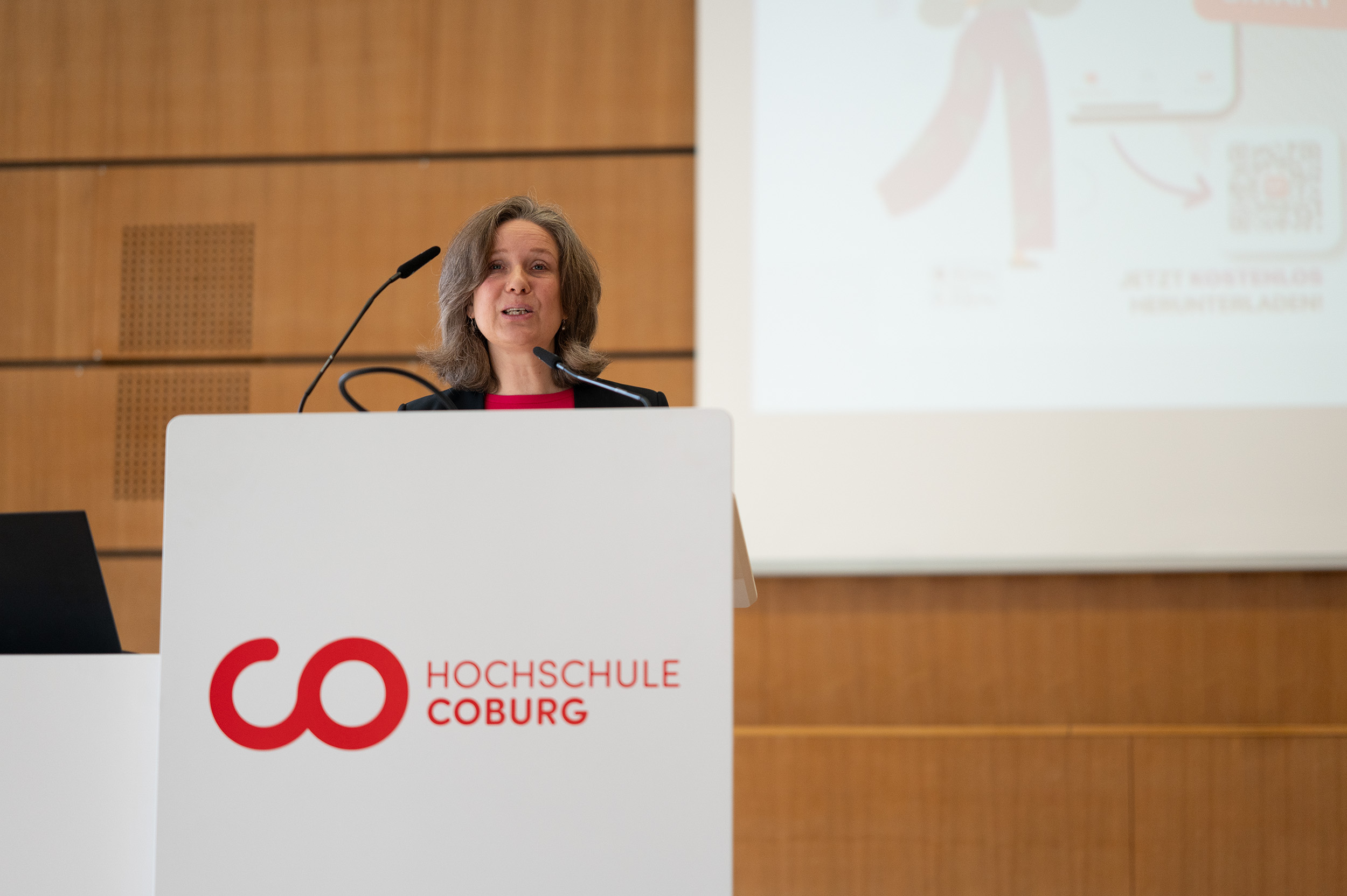 A woman stands at a podium speaking into a microphone. The podium has the logo and text "Hochschule Coburg." A presentation slide is partially visible in the background. The setting appears to be a formal event or lecture. at Hochschule Coburg