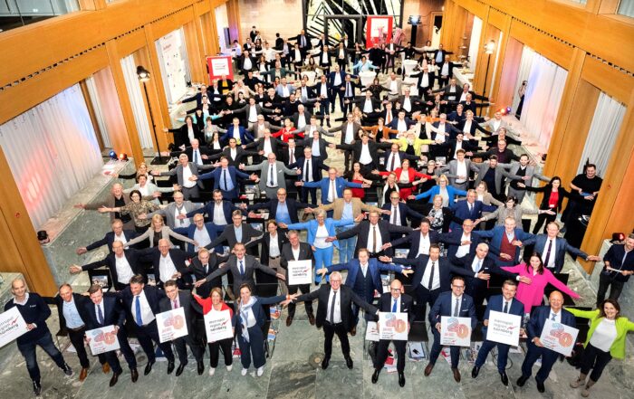 Eine große Gruppe von Menschen in Businesskleidung steht in mehreren Reihen und streckt die Arme aus, um Abstand zu halten. Einige halten Schilder mit der Aufschrift „30“ hoch. Der großzügige Saal, geschmückt mit Holzakzenten und roten Bannern, feiert „20 Jahre“ Verbundenheit in der lebendigen Metropolregion Nürnberg. at Hochschule Coburg