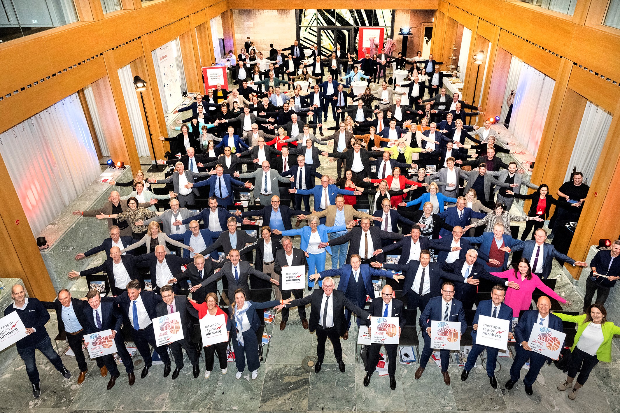 Eine große Gruppe von Menschen in Businesskleidung steht in mehreren Reihen und streckt die Arme aus, um Abstand zu halten. Einige halten Schilder mit der Aufschrift „30“ hoch. Der großzügige Saal, geschmückt mit Holzakzenten und roten Bannern, feiert „20 Jahre“ Verbundenheit in der lebendigen Metropolregion Nürnberg. at Hochschule Coburg