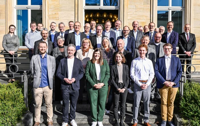 Eine Gruppe von 33 Personen posiert auf den Stufen eines Gebäudes mit großen Fenstern für ein Foto und feiert ihre kürzlich verliehene Auszeichnung. In Business- oder Abendgarderobe stehen sie vor einer Kulisse aus Steinfassade und Grün, halten stolz ihren IHK-Preis in den Händen und lächeln in die Kamera. at Hochschule Coburg