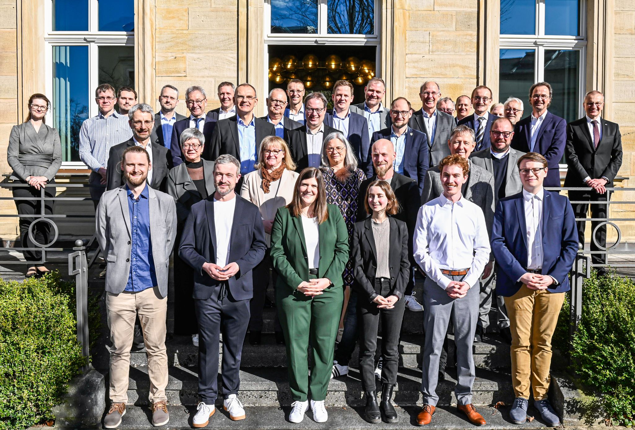 Eine Gruppe von 33 Personen posiert auf den Stufen eines Gebäudes mit großen Fenstern für ein Foto und feiert ihre kürzlich verliehene Auszeichnung. In Business- oder Abendgarderobe stehen sie vor einer Kulisse aus Steinfassade und Grün, halten stolz ihren IHK-Preis in den Händen und lächeln in die Kamera. at Hochschule Coburg