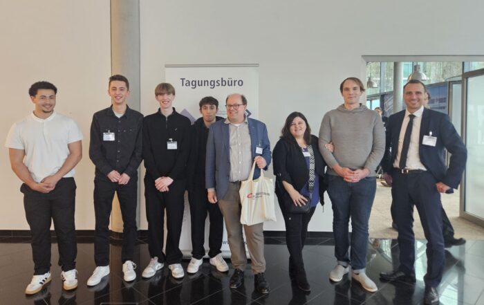Eine Gruppe von acht Personen steht drinnen vor einer Wand mit einem Schild mit der Aufschrift „Tagungsbüro“, was darauf schließen lässt, dass sie an einer Versicherungskonferenz teilnehmen. Sie tragen legere und halbformelle Kleidung, halten Konferenzausweise und Tragetaschen in den Händen und sind bereit, über Risiken und Nachhaltigkeit zu diskutieren. at Hochschule Coburg