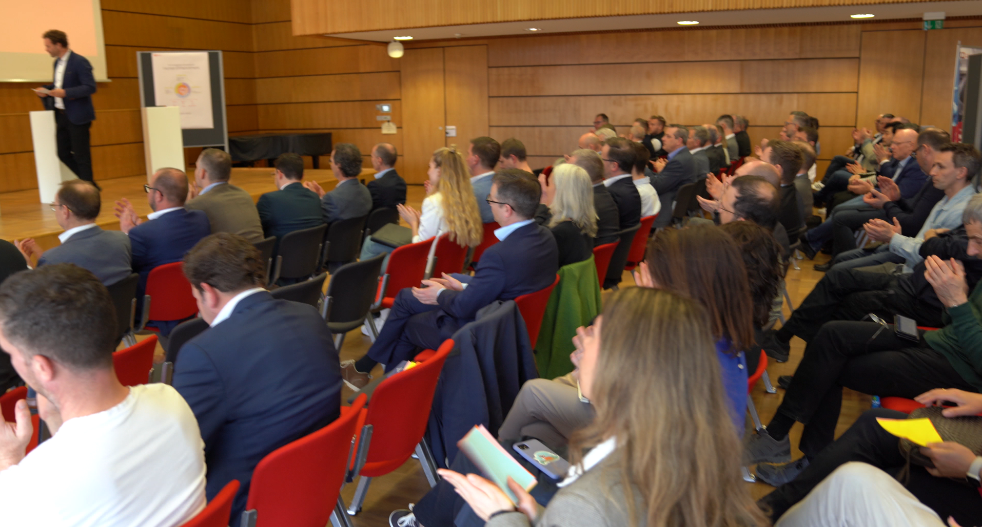 Das Publikum im Konferenzraum, das auf roten Stühlen sitzt und einem Redner am linken Podium gegenübersteht, spiegelt eine regionale Initiative zur Förderung wider. Zwischen den Holzwänden und der hohen Decke applaudieren mehrere Teilnehmer oder machen sich Notizen. at Hochschule Coburg
