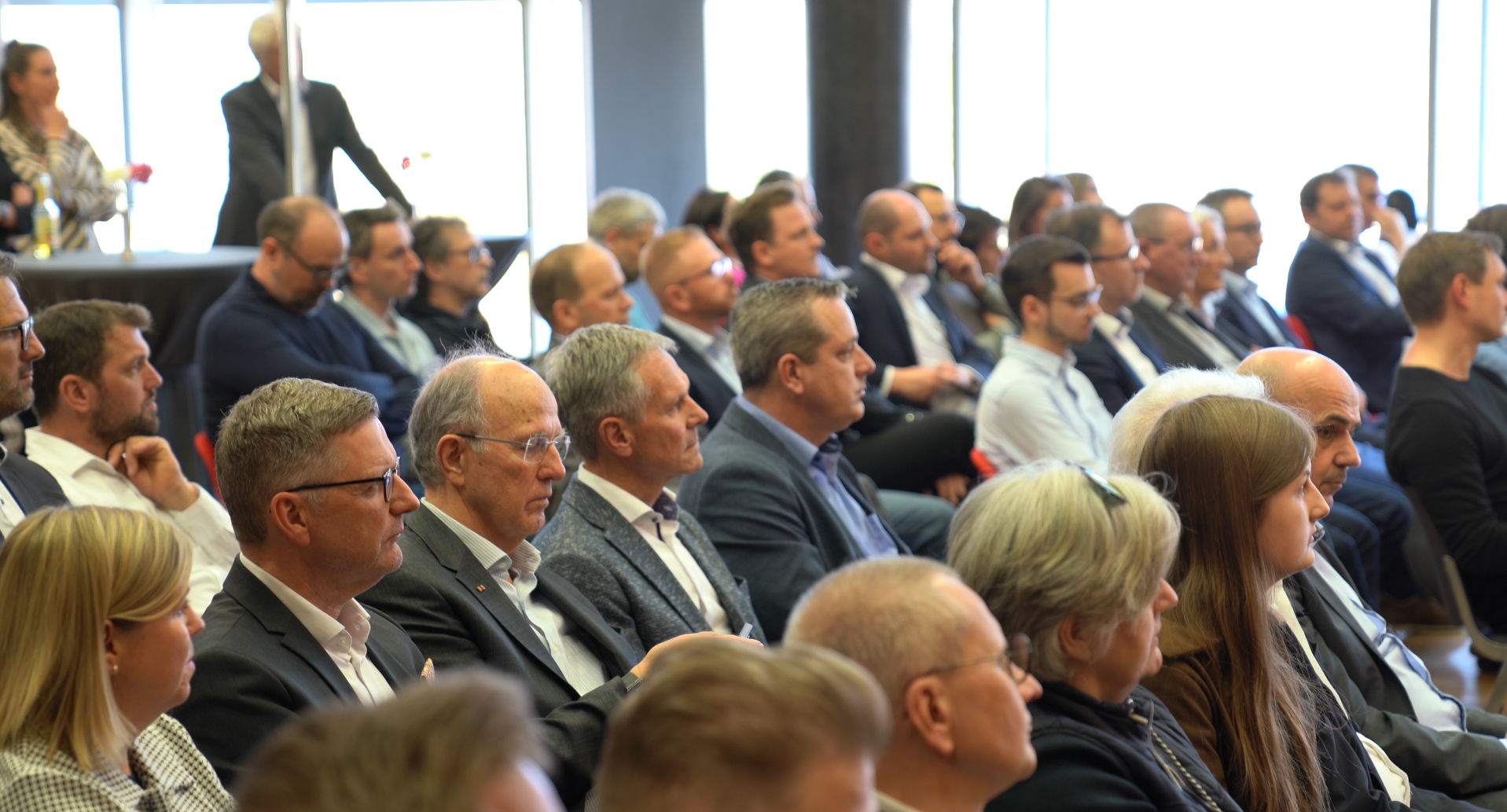 Ein Publikum aus Fachleuten unterschiedlichster Herkunft hört während einer Konferenz in einem hell erleuchteten Raum aufmerksam zu. Viele Teilnehmer tragen Anzüge und Brillen, halten Notizbücher und Papiere in der Hand und verkörpern damit bemerkenswerten Fortschritt. Im Hintergrund stehen einige Personen an den Fenstern. at Hochschule Coburg
