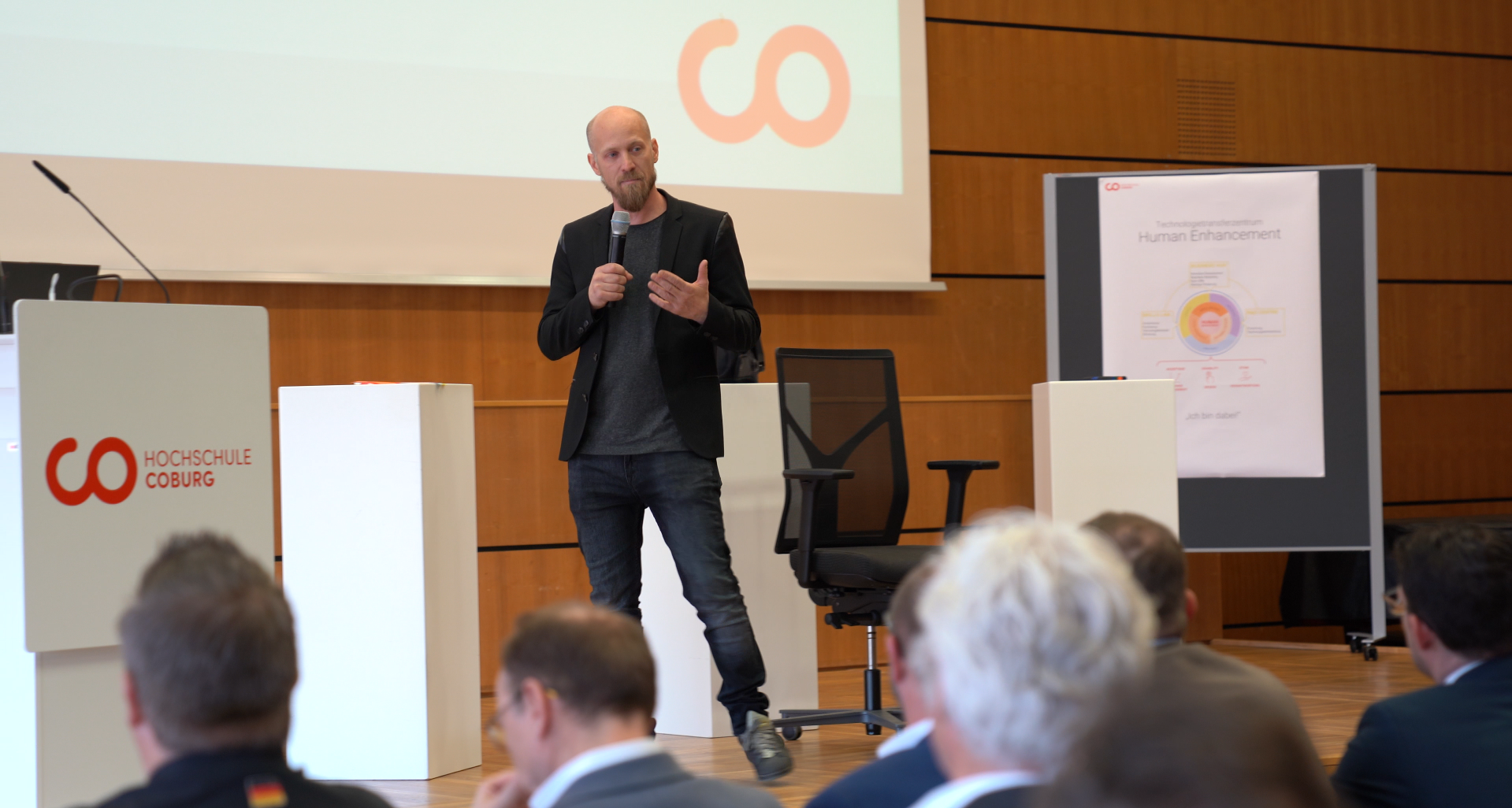 Ein Mann in schwarzem Blazer und Jeans spricht auf einer Bühne in ein Mikrofon. Hinter ihm ist ein großer Bildschirm mit einem Logo und einem Plakat mit der Aufschrift „Human Enhancement Initiative“ zu sehen. Im Vordergrund sitzen mehrere Personen, dem Sprecher zugewandt. at Hochschule Coburg