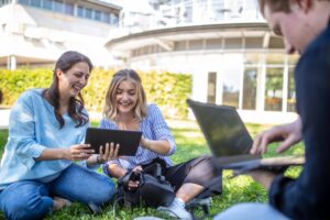 Three young adults sit on grass outside a modern building, two women smiling and looking at a tablet, while another person works on a laptop. A virtual reality headset sits on the ground beside them. at Hochschule Coburg