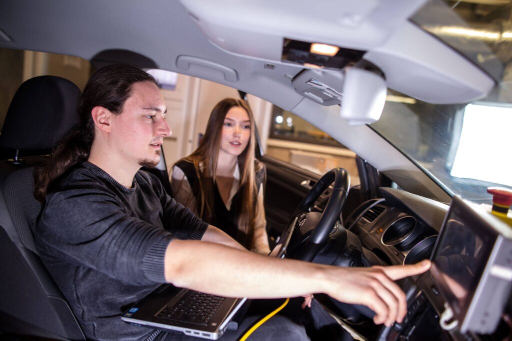 A man and a woman sit in a car, with the man using a laptop and touching a screen on the dashboard while the woman observes. The setting appears to be a garage or lab environment. at Hochschule Coburg