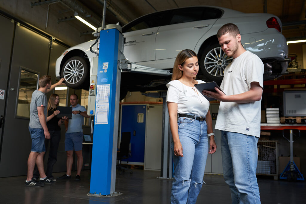 Two people stand in a garage looking at a tablet, with a car lifted on a hydraulic lift behind them. In the background, two others converse and use a tablet. All are casually dressed. at Hochschule Coburg