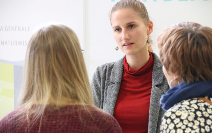 Eine Frau in grauem Blazer und rotem Rollkragenpullover unterhält sich mit zwei Personen über die Hochschule Schnuppertage. Die entspannte Atmosphäre im Innenraum bietet die perfekte Kulisse für ihr Gespräch. Die beiden hören aufmerksam zu und blicken von der Kamera weg. at Hochschule Coburg