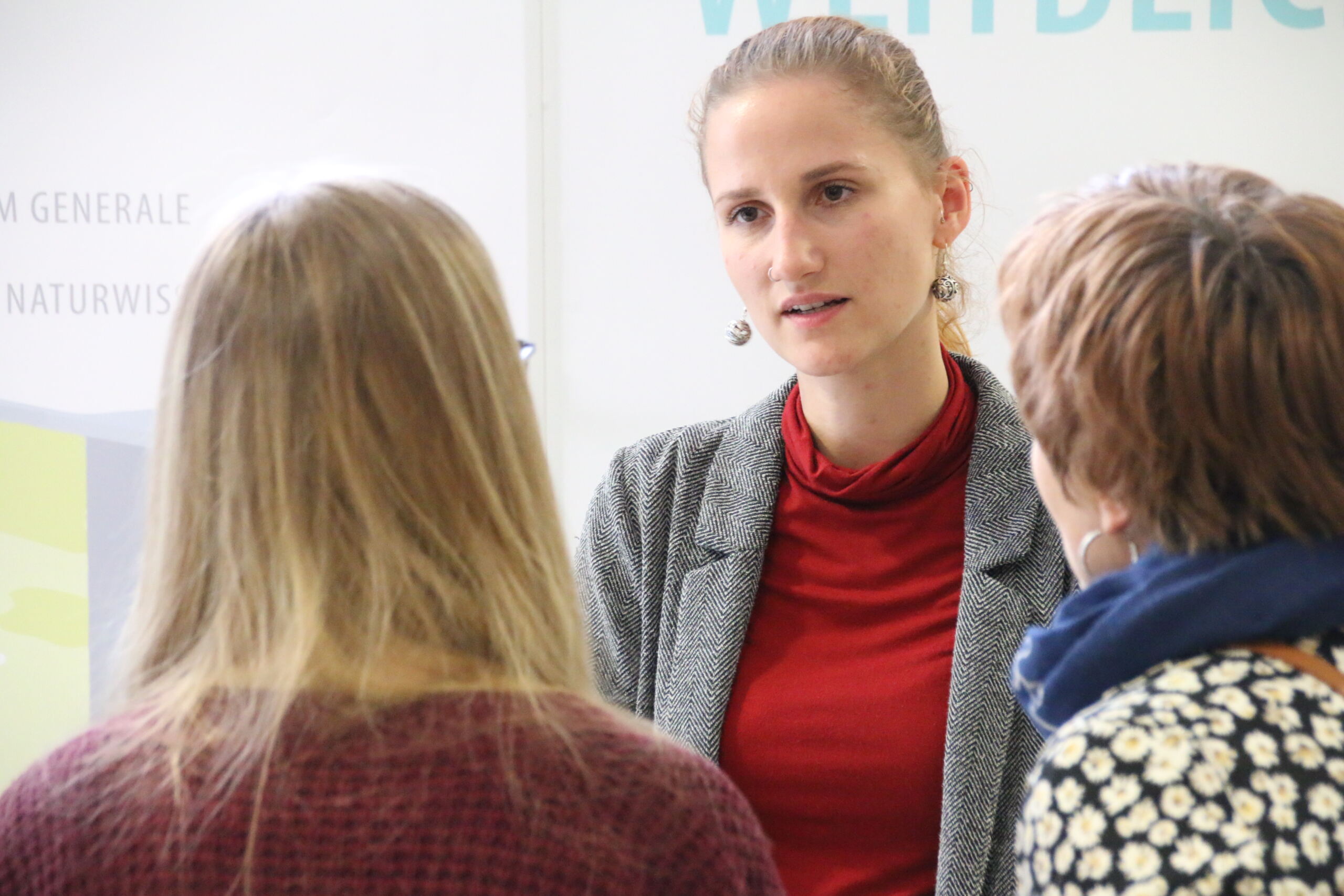 Eine Frau in grauem Blazer und rotem Rollkragenpullover unterhält sich mit zwei Personen über die Hochschule Schnuppertage. Die entspannte Atmosphäre im Innenraum bietet die perfekte Kulisse für ihr Gespräch. Die beiden hören aufmerksam zu und blicken von der Kamera weg. at Hochschule Coburg