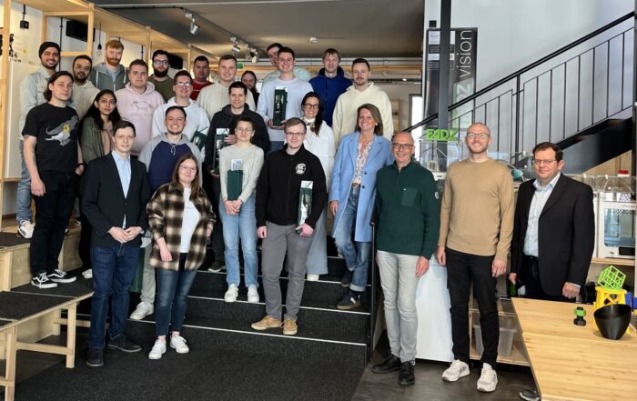 Eine Gruppe von Masterstudierenden posiert gemeinsam in einem modernen Gebäude mit Industriedekor in Lichtenfels. Sie stehen vor einer Treppe, umgeben von Regalen und Tischen. Einige halten Getränke in den Händen, während Informationsplakate zur lebendigen Atmosphäre beitragen. at Hochschule Coburg