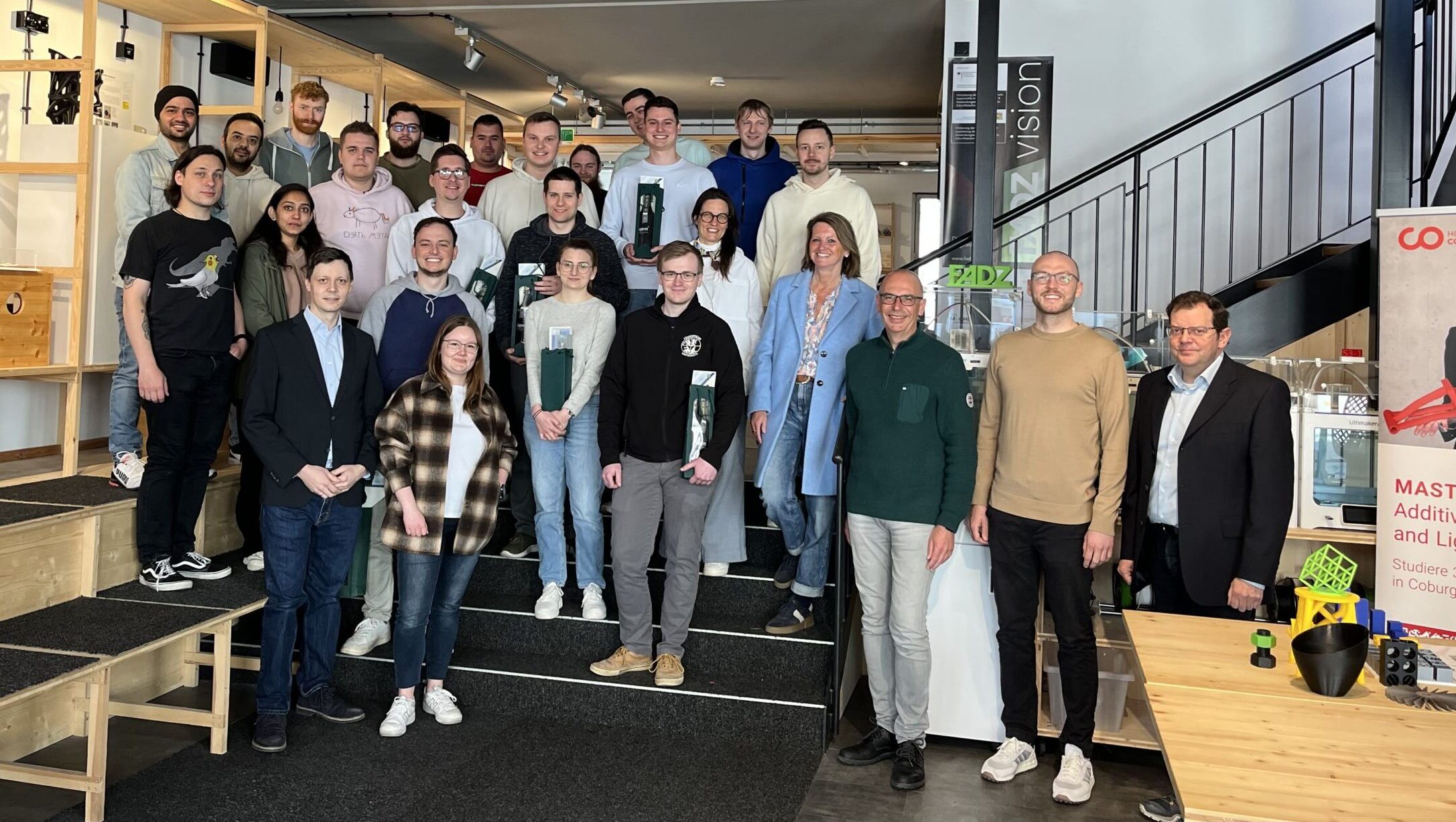 Eine Gruppe von Masterstudierenden posiert gemeinsam in einem modernen Gebäude mit Industriedekor in Lichtenfels. Sie stehen vor einer Treppe, umgeben von Regalen und Tischen. Einige halten Getränke in den Händen, während Informationsplakate zur lebendigen Atmosphäre beitragen. at Hochschule Coburg