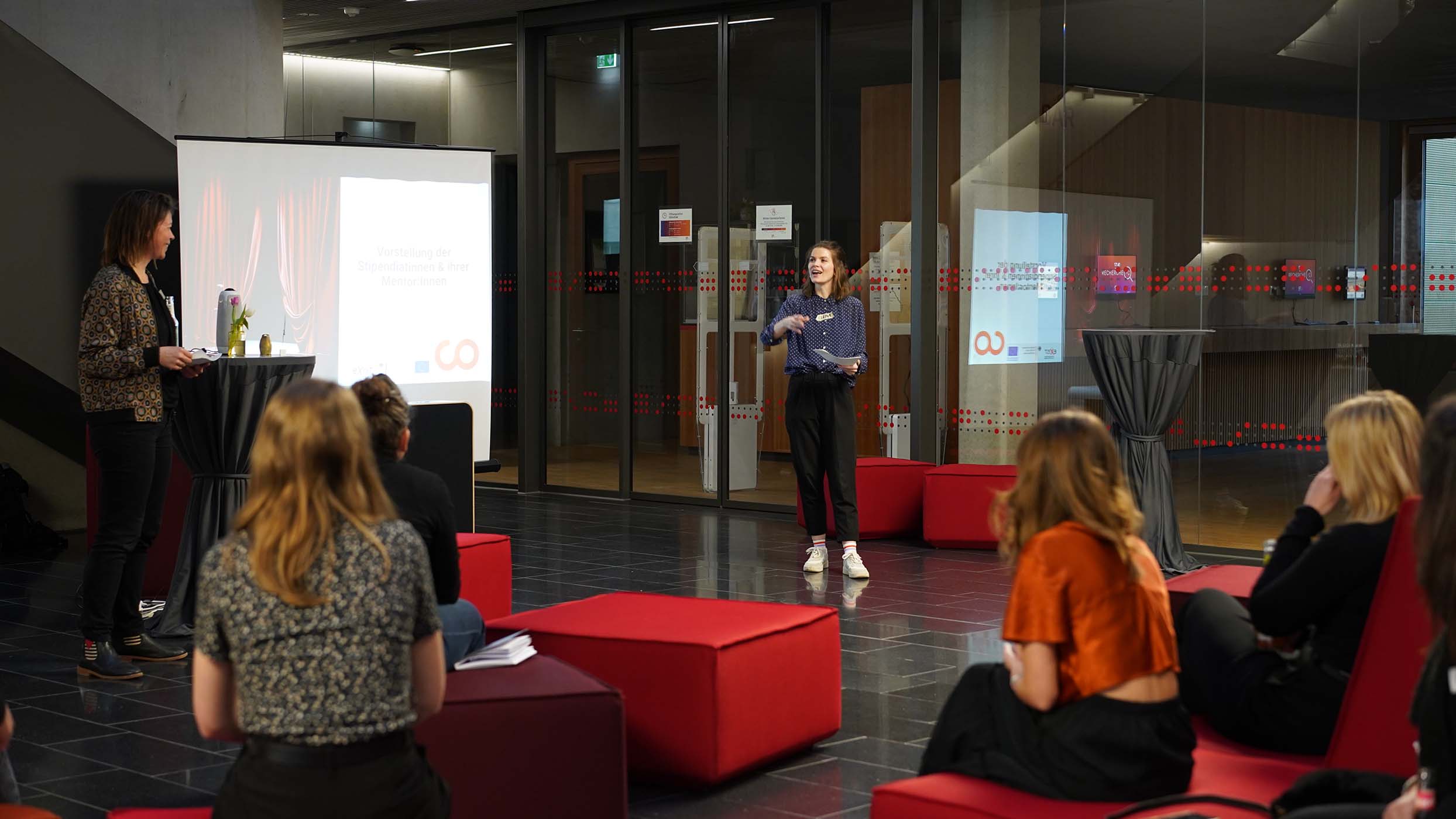 Ein Redner im blauen Hemd präsentiert bei einer Indoor-Veranstaltung, bei der Networking-Gespräche in der Luft liegen. Auf einem Bildschirm wird eine Folie angezeigt, während die Teilnehmer aufmerksam zuhören. Der moderne Raum mit seinen Glastüren und dekorativen roten Elementen bildet die ideale Kulisse für diesen spannenden Ideenaustausch. at Hochschule Coburg