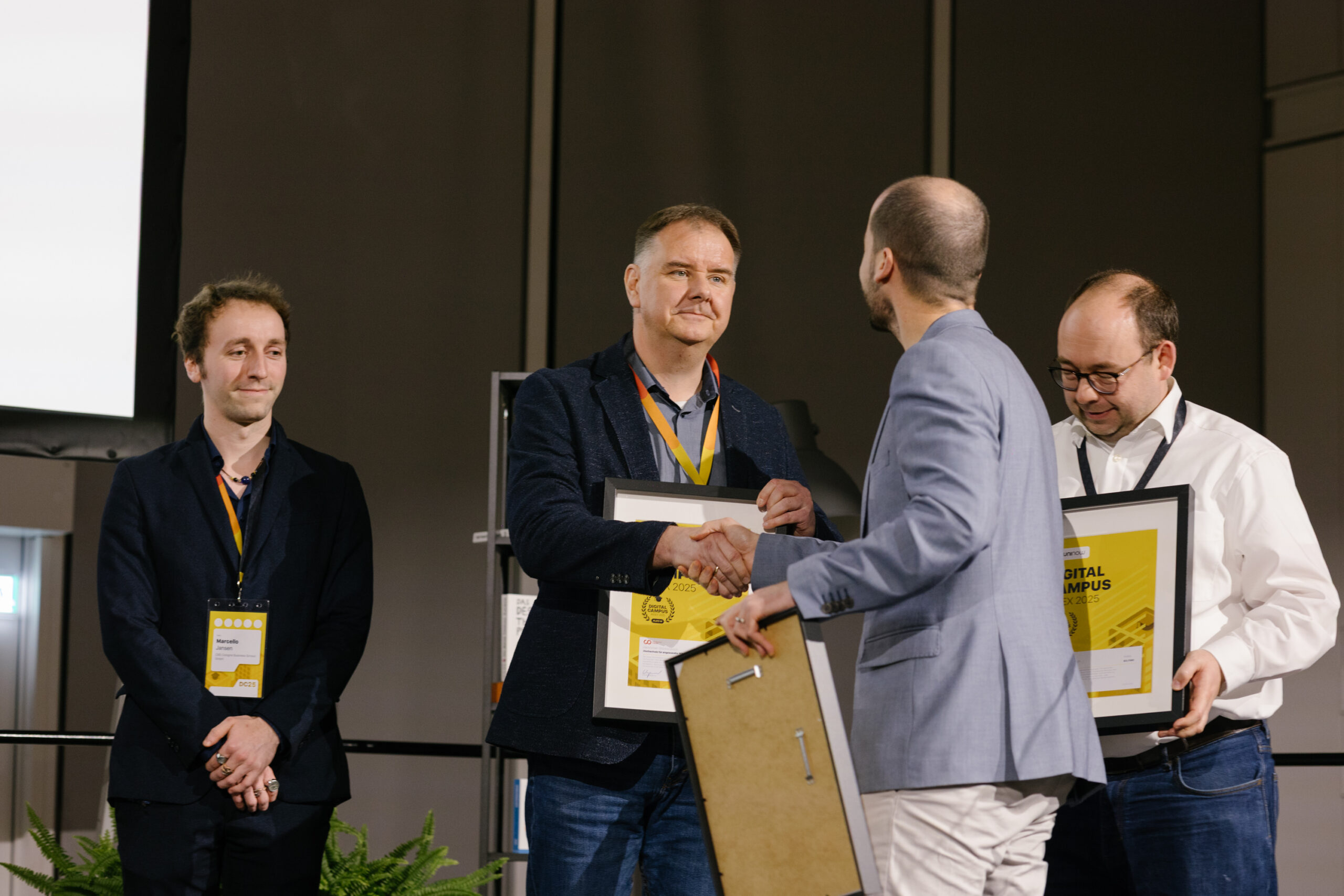 Vier Männer stehen auf der Bühne, zwei schütteln Hände und halten gerahmte Urkunden für den DigitalCampus-Award 2025 in den Händen. Ein weiterer Mann steht lächelnd links und ein vierter – stellvertretend für die Hochschule Coburg – hält ebenfalls stolz eine Urkunde in den Händen. at Hochschule Coburg