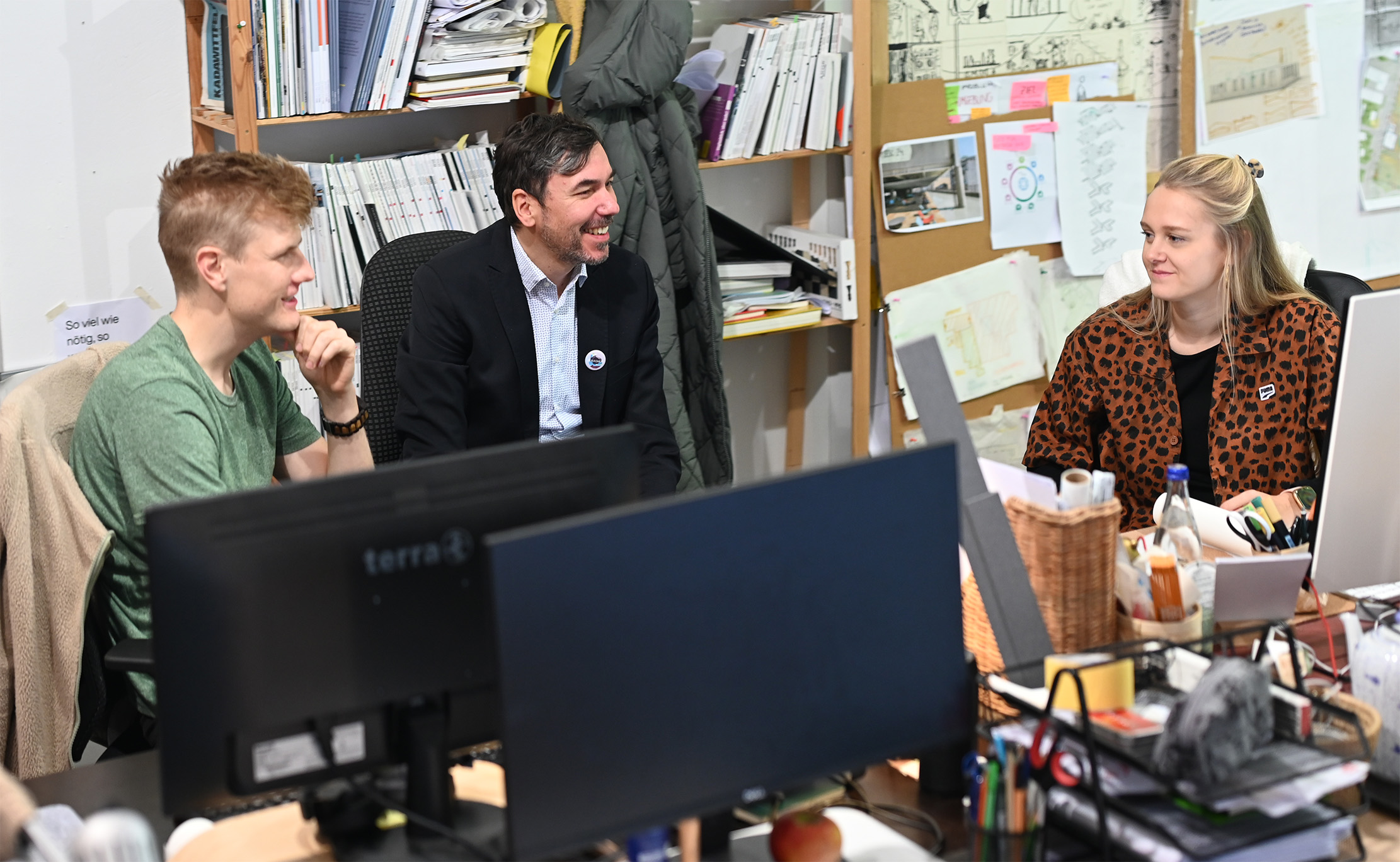 Drei Personen unterhalten sich in einem Büro. Zwei Männer und eine Frau lächeln an ihren Schreibtischen mit Computern, umgeben von Regalen voller Bücher und Dokumente. Die Atmosphäre ist entspannt und kollegial. at Hochschule Coburg