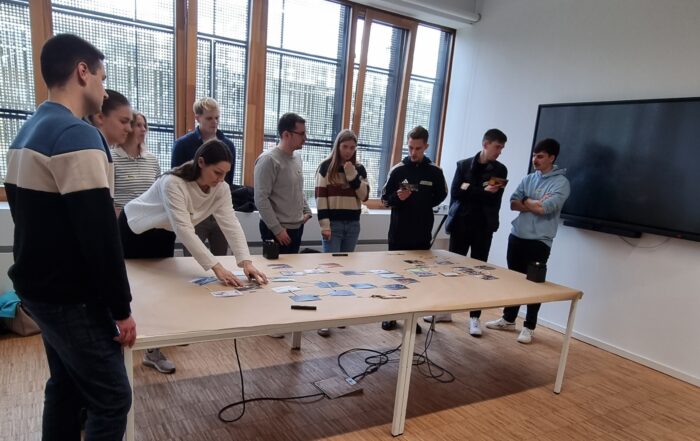 Eine Gruppe von Menschen steht um einen mit Papieren und Karten bedeckten Tisch und diskutiert über das Klimapuzzle. In einem hellen Raum mit großen Fenstern und einem an der Wand montierten Bildschirm arbeiten sie eifrig zusammen, entschlossen, etwas gegen die Klimakrise zu unternehmen. at Hochschule Coburg