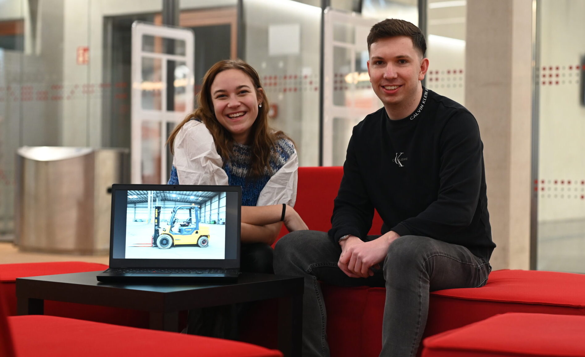 Zwei junge Erwachsene sitzen auf roten Stühlen in einer modernen Lounge und lächeln in die Kamera. Vor ihnen zeigt ein Laptop ein Fahrzeugbild – möglicherweise aus dem Bereich Logistik oder Wirtschaftsinformatik. Glastüren und modernes Dekor runden die Szene ab. at Hochschule Coburg