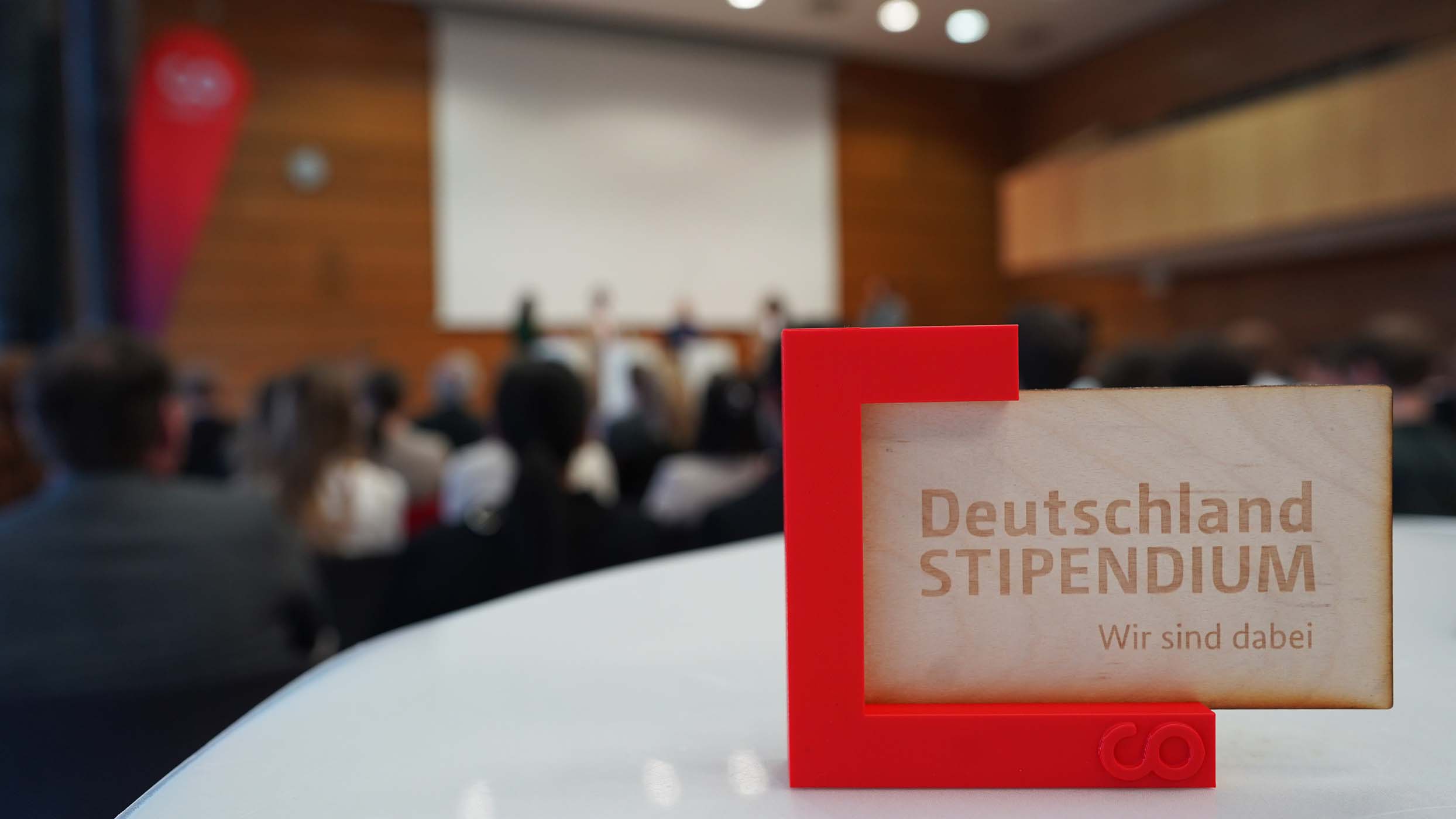 Ein rot-weißes Schild mit der Aufschrift „Deutschland STIPENDIUM Wir sind dabei“ steht im Fokus auf einem Tisch. Im unscharfen Hintergrund blicken Studierende aufmerksam auf eine Projektionsfläche in einem Hörsaal, unterstützt von engagierten Förderern, die an die Förderung zukünftiger Talente glauben. at Hochschule Coburg
