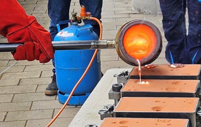 Eine Person demonstriert Handwerkstechnik, indem sie geschmolzenes Metall in rechteckige Formen auf einem Tisch im Freien gießt, während drei Beobachter aufmerksam zuschauen. Im Hintergrund ragt ein blauer Gastank hervor, der dieser improvisierten Designausbildung einen industriellen Touch verleiht. at Hochschule Coburg