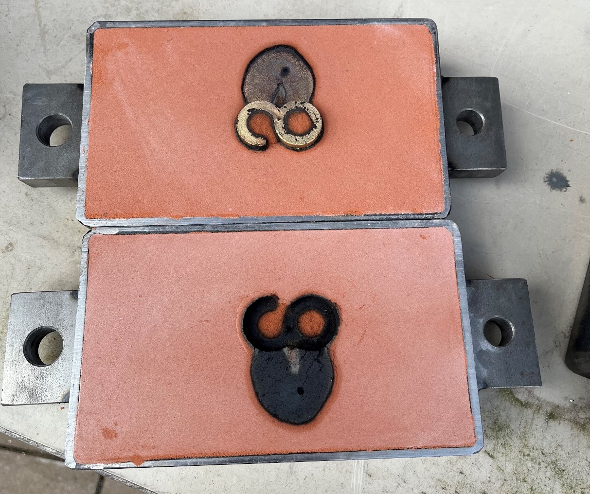 Zwei rechteckige Tonplatten mit Metallgriffen, die jeweils ein schwarz gebranntes Muster aus Schleifen und Kreisen auf einer rosa-orangen Oberfläche zeigen, unterstreichen die in der Designausbildung vermittelte Handwerkskunst. Die Platten liegen perfekt auf einem hellgrauen Hintergrund und spiegeln die sorgfältige Handwerkstechnik wider. at Hochschule Coburg