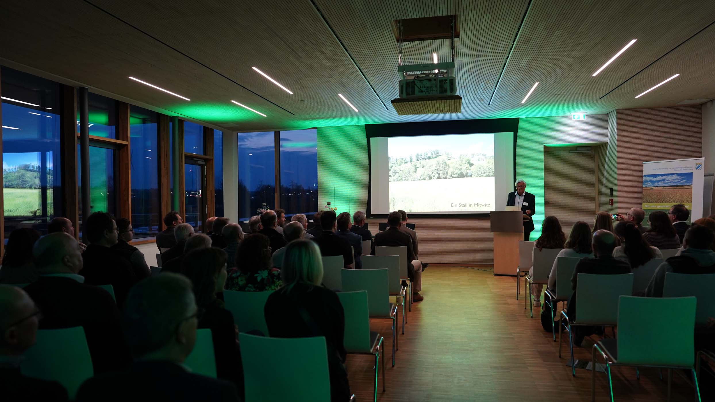 In einem modernen Konferenzraum präsentiert ein Redner vor sitzenden Teilnehmern. Der in Grüntönen gehaltene Raum bietet durch große Fenster einen Blick auf die Abendlandschaft von Münchberg. Auf der Präsentationsleinwand verweben studentische Entwürfe Architektur und Landschaft auf kreative Weise. at Hochschule Coburg