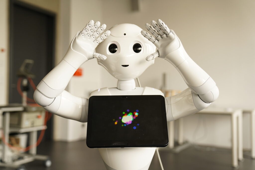 A white humanoid robot holds its hands near its head, appearing playful. It has a tablet on its chest displaying colorful dots. The background shows a bright, modern room with lab equipment and tables. at Hochschule Coburg