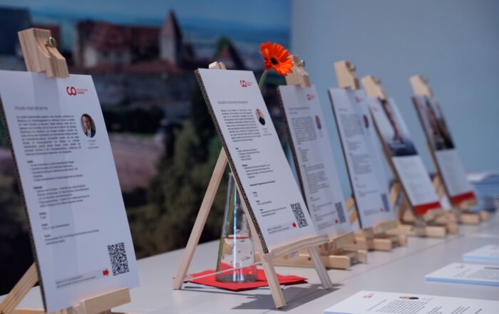 Verschiedene Informationsbroschüren, ähnlich einem sorgfältig gestalteten Entwurf, liegen auf Staffeleien in einer Reihe auf einem Tisch. Der Fokus liegt auf den Broschüren, die jeweils Text und QR-Codes enthalten. Im Hintergrund, etwa in der Mitte des Tisches, ist eine einzelne orangefarbene Blume zu sehen. at Hochschule Coburg