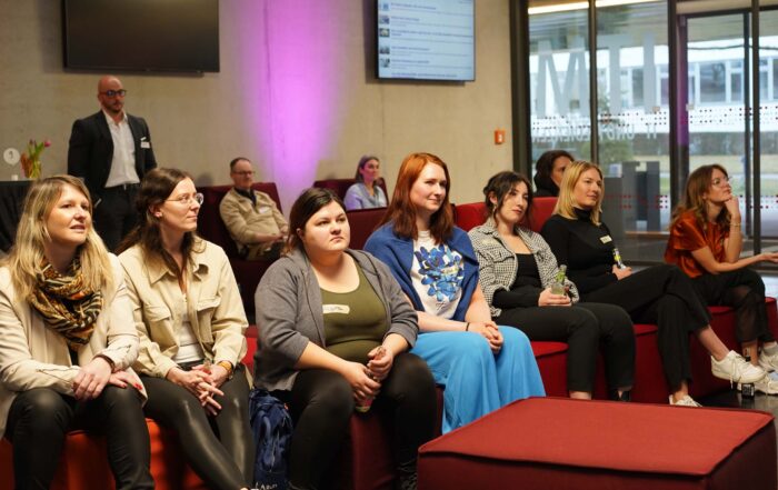 In einem modernen Interieur sitzt eine Gruppe von Menschen aufmerksam auf roten Sofas und pflegt angeregtes Networking. Einige stehen oder sitzen im Hintergrund, während ein großer Bildschirm den sanft beleuchteten Raum mit rosa und natürlichem Licht ziert. at Hochschule Coburg