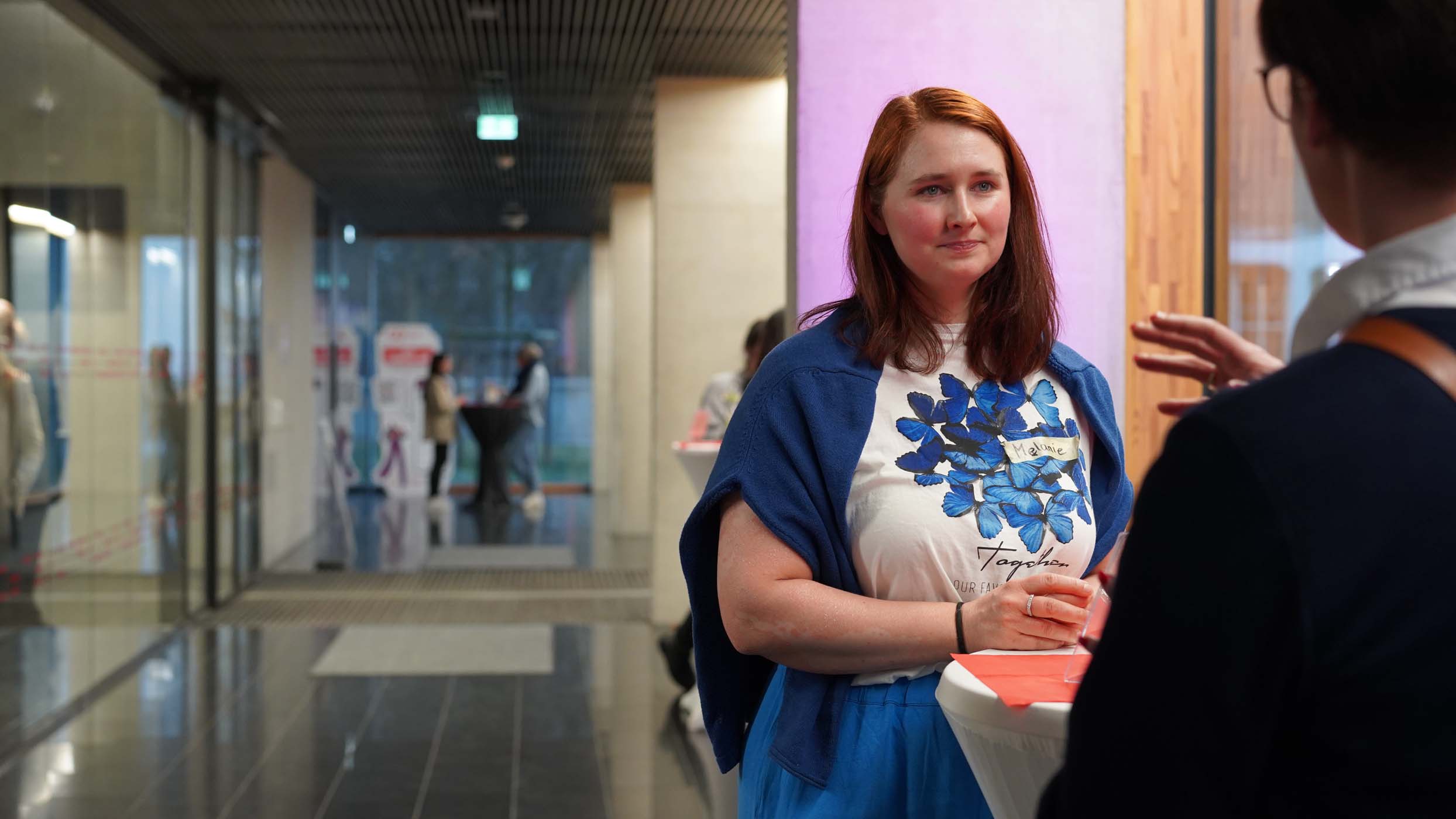 Eine Frau steht im Flur einer foundHER-Veranstaltung. Sie trägt ein weißes Hemd mit blauem Blumenmuster und eine blaue Strickjacke. Aufmerksam lauscht sie den gestikulierenden Blicken einer Person. Der Flur, perfekt für die Auftaktveranstaltung, besticht durch Glaswände, einen reflektierenden Boden und mehrere Personen im Hintergrund. at Hochschule Coburg
