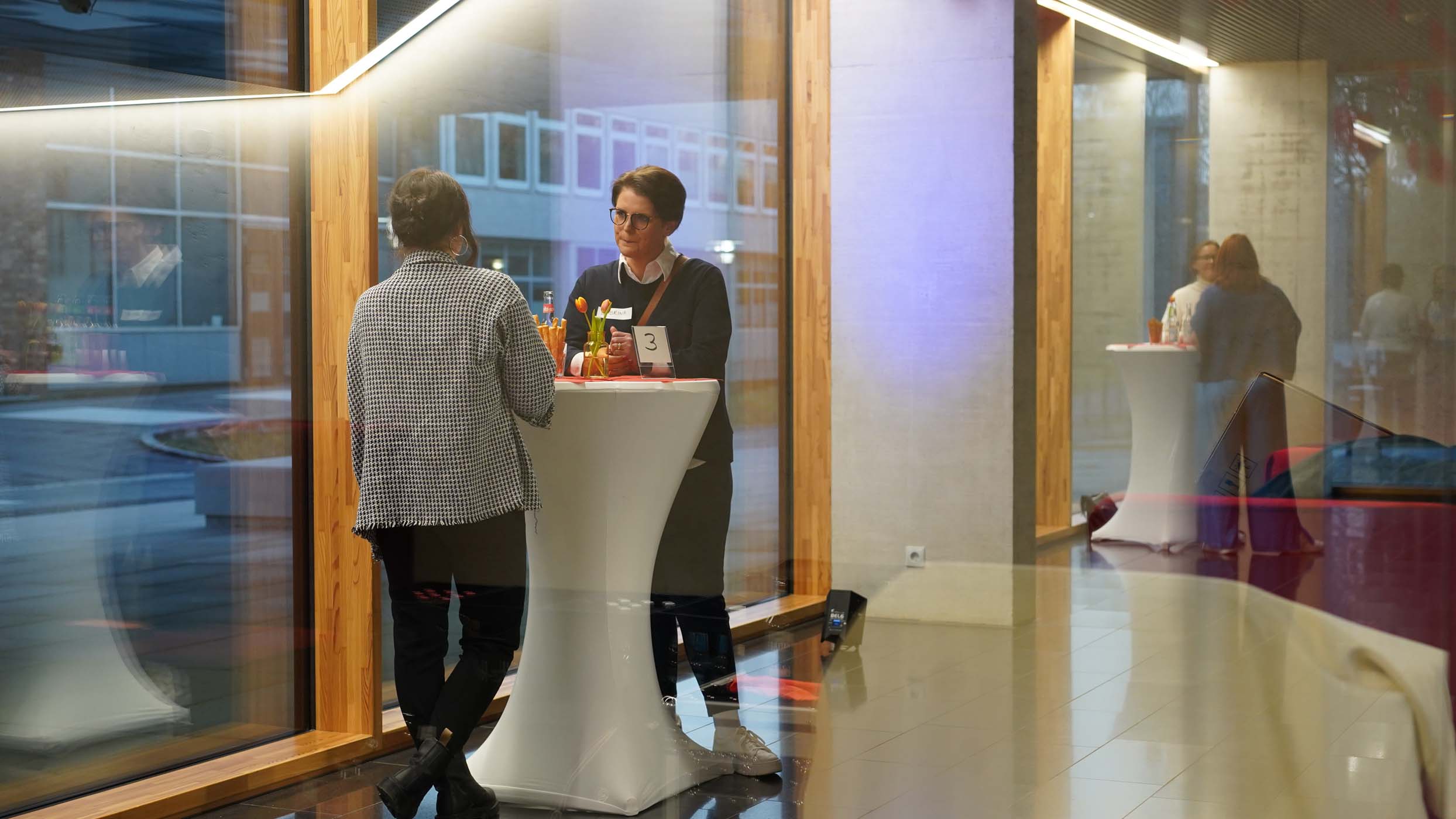 Zwei Personen stehen an einem hohen runden Tisch und unterhalten sich zum Speed-Dating in einer modernen Lobby mit Glaswänden. Der Raum ist sanft beleuchtet, mit Spiegelungen auf dem glänzenden Boden. Im Hintergrund ist eine weitere Person an einem ähnlichen Tisch zu sehen, die sich in Networking-Gespräche vertieft. at Hochschule Coburg
