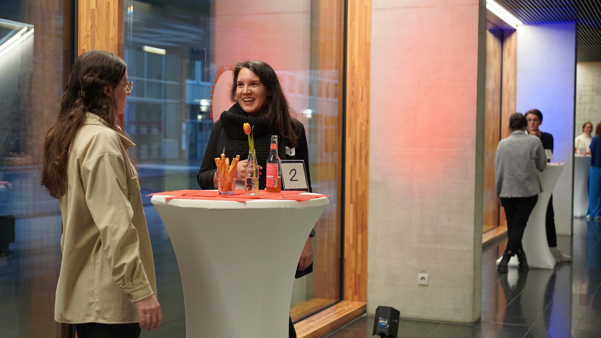 Zwei Personen stehen mit Getränken und einer Tulpe im Glas an einem Stehtisch und knüpfen angeregtes Networking. Eine Person im schwarzen Mantel lächelt, während die andere, in einer beigen Jacke, aufmerksam zuhört. Die anderen Teilnehmer mischen sich in dem modernen, gut beleuchteten Raum, der die Essenz der foundHER-Veranstaltungen einfängt. at Hochschule Coburg
