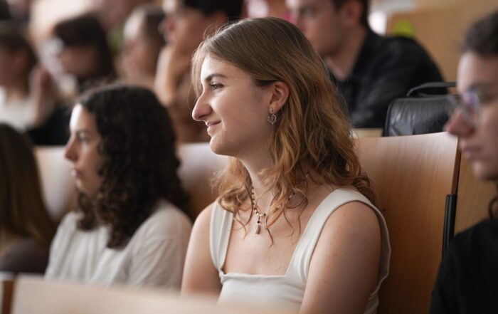 Eine junge Frau mit welligem hellbraunem Haar und weißem Oberteil sitzt im Hörsaal des Studiums Coburg und hört aufmerksam zu. Um sie herum sitzen andere Studierende, die das lebendige Studentenleben Coburgs genießen und sich auf die Rednerin konzentrieren. at Hochschule Coburg