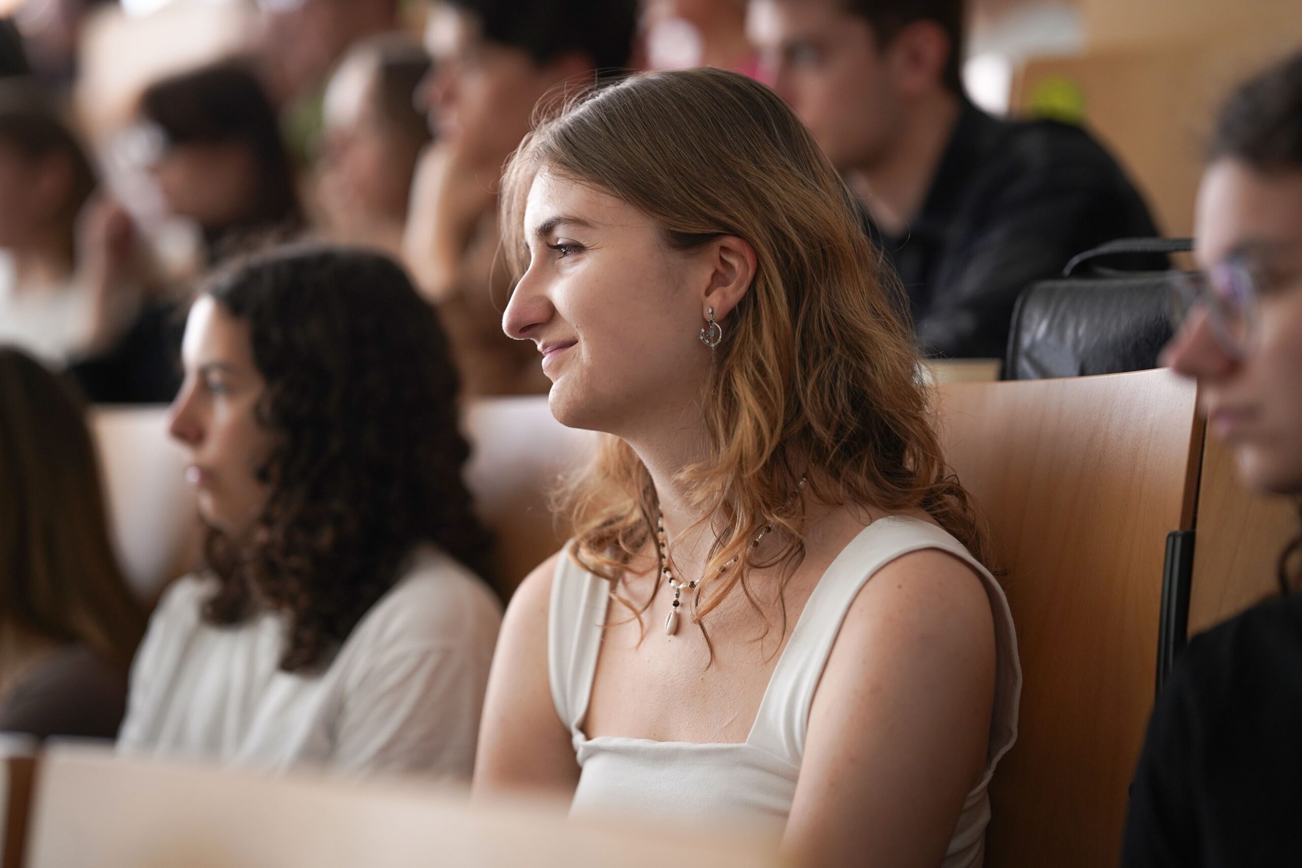 Eine junge Frau mit welligem hellbraunem Haar und weißem Oberteil sitzt im Hörsaal des Studiums Coburg und hört aufmerksam zu. Um sie herum sitzen andere Studierende, die das lebendige Studentenleben Coburgs genießen und sich auf die Rednerin konzentrieren. at Hochschule Coburg