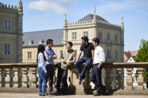 Five young adults stand and sit on a stone balcony in the student city Coburg, talking and smiling, with a historic European-style building and blue sky in the background. They appear relaxed and engaged in conversation. at Hochschule Coburg