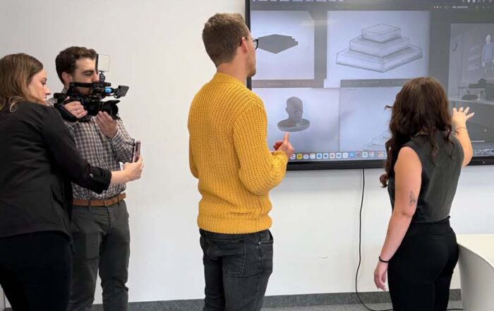Vier Personen befinden sich in einem Büro. Zwei filmen, während zwei weitere an einem großen Bildschirm stehen, auf dem 3D-Modelle gezeigt werden – eine spannende Szene, die die praxisorientierte Ausbildung in Wirtschaftsinformatik für die digitale Zukunft unterstreicht. at Hochschule Coburg