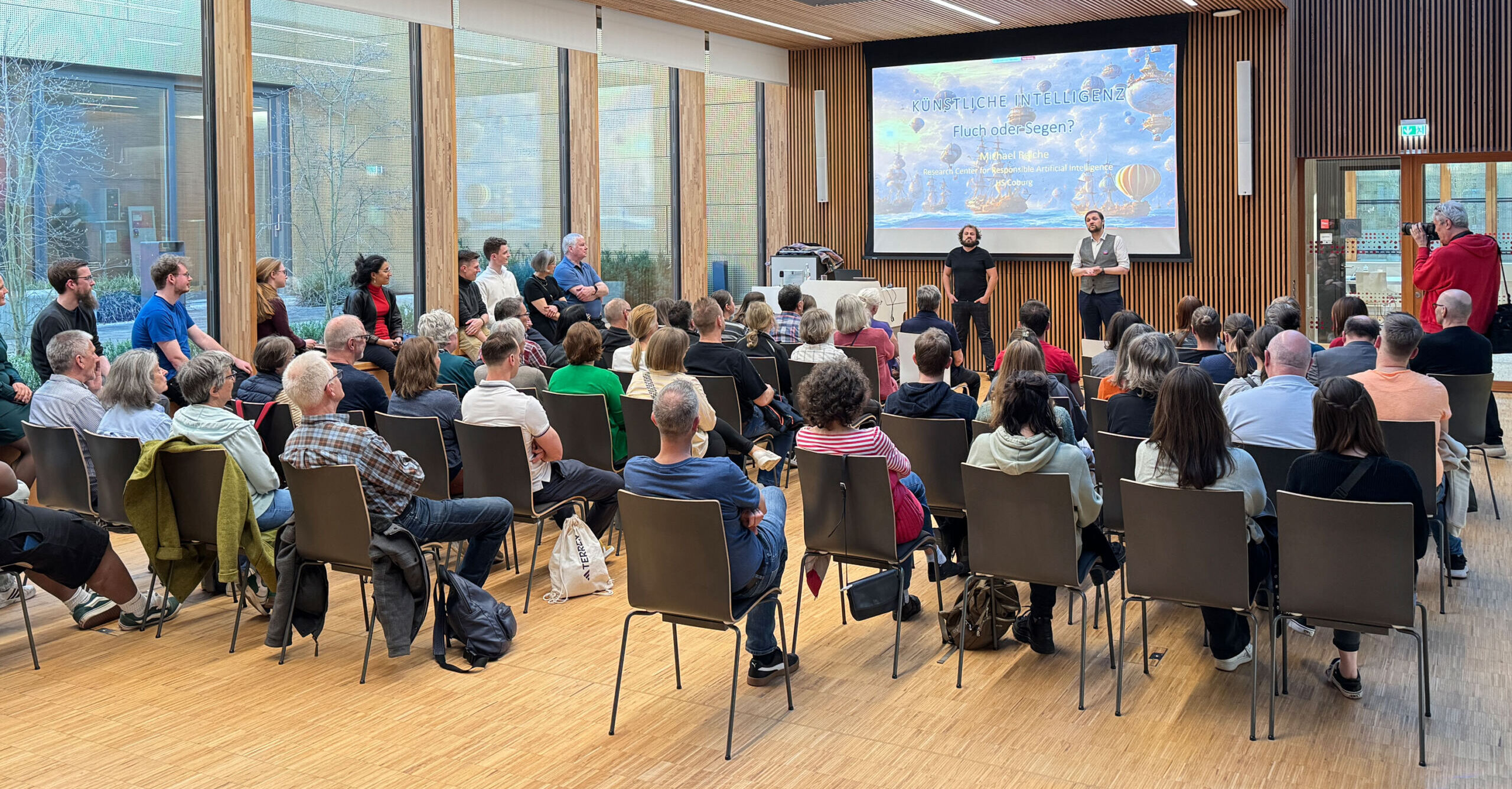 Eine große Gruppe von Menschen sitzt in einem modernen Konferenzraum mit Holzwänden und großen Fenstern und lauscht aufmerksam den Vorträgen zweier Redner vor einer Leinwand mit einem farbenfrohen Bild. Diese Veranstaltung in Coburg ist ein weiterer Erfolg der Nacht der Bibliotheken und begeistert alle Teilnehmer mit spannenden Inhalten. at Hochschule Coburg