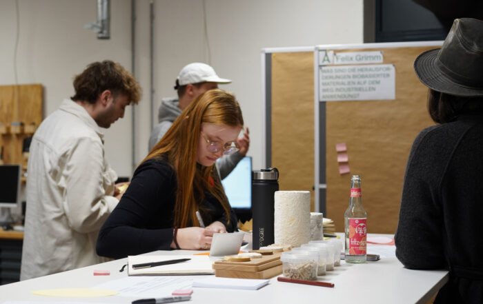 Eine Gruppe von Menschen sitzt in einem Arbeitsraum an einem Tisch mit Notizen und Getränken und diskutiert über Entwurfsstrategien für eine nachhaltige Zukunft. Eine Frau mit roten Haaren schreibt konzentriert, während andere in der Nähe stehen. Im Hintergrund sind eine Pinnwand mit Notizen und ein Poster zu sehen. at Hochschule Coburg