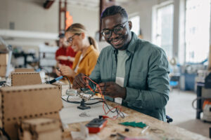 A smiling man works on an electronics project with wires at a table, while a woman in the background also works on a similar task in a well-lit, creative workshop. at Hochschule Coburg