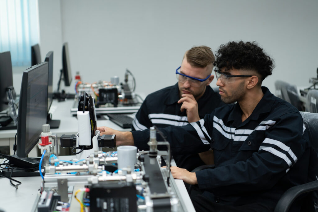 Two men wearing safety glasses and black work uniforms operate electronic equipment and computers in a laboratory or technical workspace, focusing intently on their tasks. at Hochschule Coburg