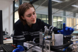 A person wearing a dark work uniform and blue gloves inspects machinery or equipment in a lab or industrial setting, with large windows and a computer in the background. at Hochschule Coburg