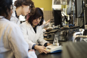 Three people in lab coats and safety goggles work together in a laboratory, using a digital device with a flask on it. Computers and lab equipment are visible on the benches around them. at Hochschule Coburg