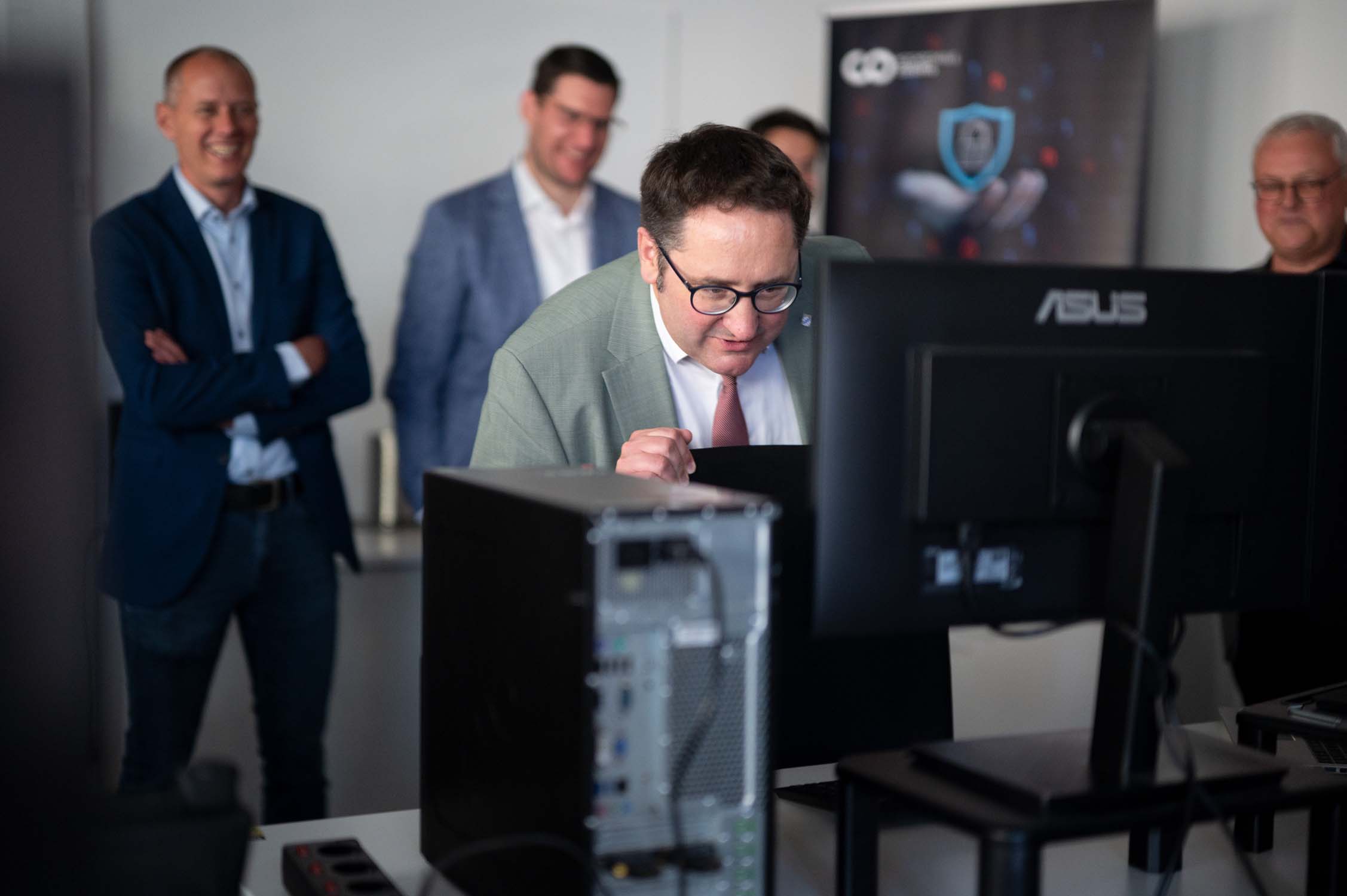 Staatssekretär Gotthardt beugt sich im Anzug nach vorn und betrachtet aufmerksam einen Computermonitor der KI-Forschung, während drei weitere Männer lächelnd hinter ihm in einem Büro stehen. Im Hintergrund ist ein verschwommenes Banner zu sehen. at Hochschule Coburg
