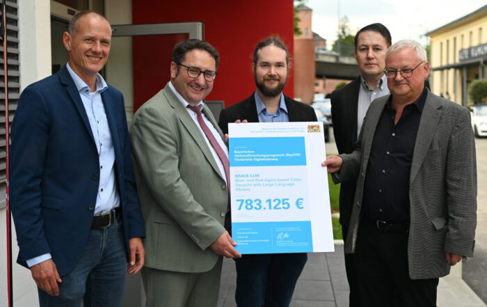 Fünf Männer in Businesskleidung stehen lächelnd vor einem Gebäude und halten einen großen Festscheck über 783.125 Euro für KI-Forschung in den Händen, wie es auf Deutsch heißt. Staatssekretär Gotthardt ist bei der von CRAI organisierten Feier anwesend. at Hochschule Coburg