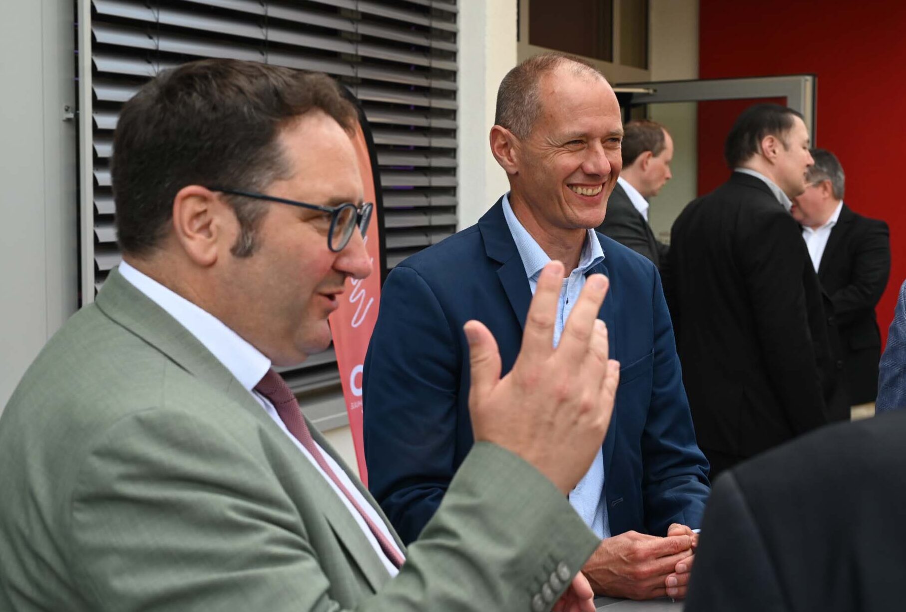 Draußen steht eine Gruppe von Männern in Businesskleidung und unterhält sich über KI-Forschung. Ein Mann im grünen Anzug gestikuliert lebhaft, ein anderer im blauen Blazer lächelt. Die Atmosphäre wirkt professionell, im Hintergrund ein modernes Gebäude. at Hochschule Coburg