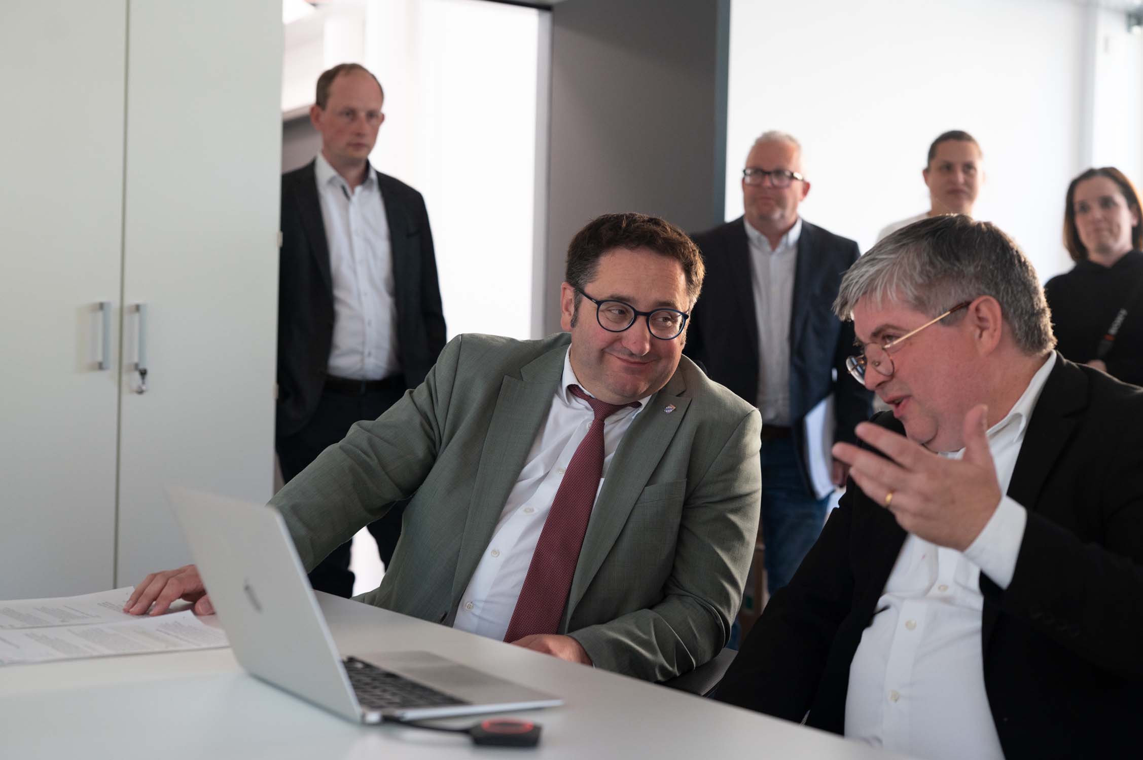 Staatssekretär Gotthardt und ein Kollege sitzen im Anzug an einem Tisch und diskutieren über KI-Forschung am Laptop, während weitere Personen im Hintergrund in einem Büro aufmerksam zuschauen. at Hochschule Coburg