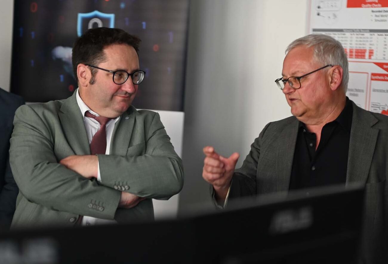 Drei Männer in Anzügen stehen in einem Büro. Zwei unterhalten sich vor einem Computerbildschirm. Einer gestikuliert, während der andere zuhört. Im Hintergrund ist eine Präsentationstafel des CRAI über KI-Forschung zu sehen. at Hochschule Coburg